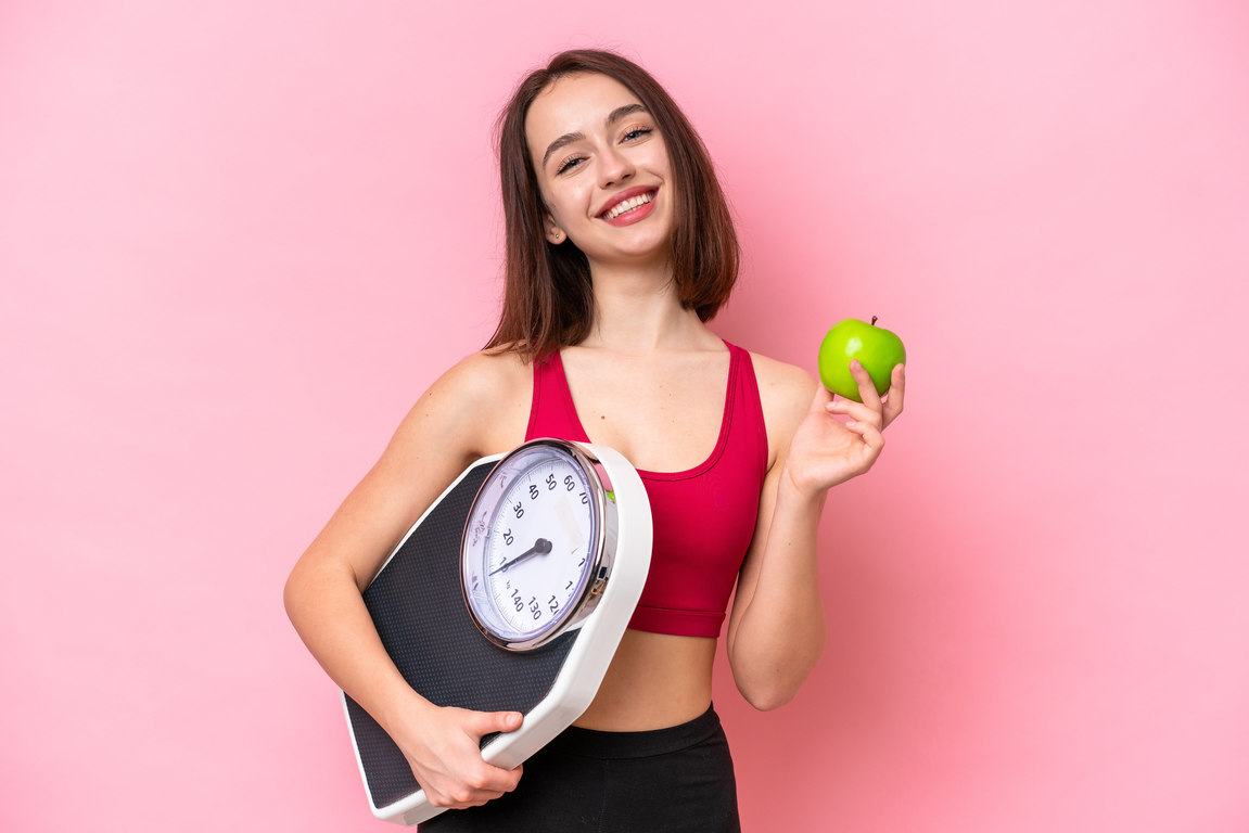 Young Ukrainian woman isolated on pink background with weighing machine and with an apple